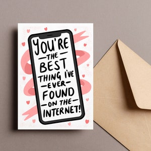 Op de afbeelding: Een wenskaart met een smartphone-afbeelding en de tekst "You're the best thing I've ever found on the internet!" in het zwart. Roze harten en een roze lintachtig ontwerp omringen de telefoon. Een bruine envelop staat rechts.