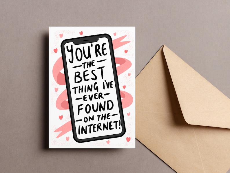 Op de afbeelding: Een wenskaart met een smartphone-afbeelding en de tekst "You're the best thing I've ever found on the internet!" in het zwart. Roze harten en een roze lintachtig ontwerp omringen de telefoon. Een bruine envelop staat rechts.