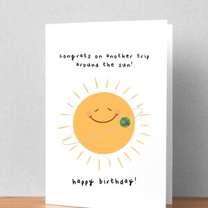 K&ouml;nnte beinhalten: Wei&szlig;e Geburtstagskarte mit einer Illustration einer l&auml;chelnden Sonne und einem Planeten in der Umlaufbahn. Der Text lautet: &bdquo;Herzlichen Gl&uuml;ckwunsch zu einer weiteren Reise um die Sonne!&ldquo; und &bdquo;Alles Gute zum Geburtstag!&ldquo; in Schwarz.