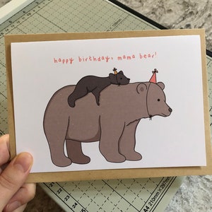 Pu&ograve; includere: Un biglietto di auguri con un bordo marrone. La carta presenta un'illustrazione di un orso bruno grande con un orso nero pi&ugrave; piccolo sulla schiena, entrambi con cappelli da festa. Il testo "happy birthday, mama bear!" &egrave; scritto in rosso.