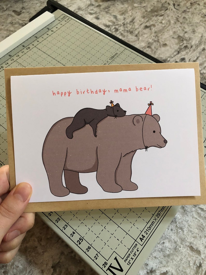 Pu&ograve; includere: Un biglietto di auguri con un bordo marrone. La carta presenta un'illustrazione di un orso bruno grande con un orso nero pi&ugrave; piccolo sulla schiena, entrambi con cappelli da festa. Il testo "happy birthday, mama bear!" &egrave; scritto in rosso.