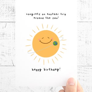 K&ouml;nnte beinhalten: Geburtstagskarte mit einer Illustration einer l&auml;chelnden Sonne mit gelben Strahlen und einer kleinen Erdkugel. Der Text lautet: "Herzlichen Gl&uuml;ckwunsch zu einer weiteren Sonnenumrundung!" und "Alles Gute zum Geburtstag!"