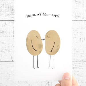 May include: A greeting card with the text "YOU'RE MY BEST SPUD!" features two cartoon potatoes with arms and legs. The potatoes are light brown with darker spots and are smiling. The card is held by a hand.