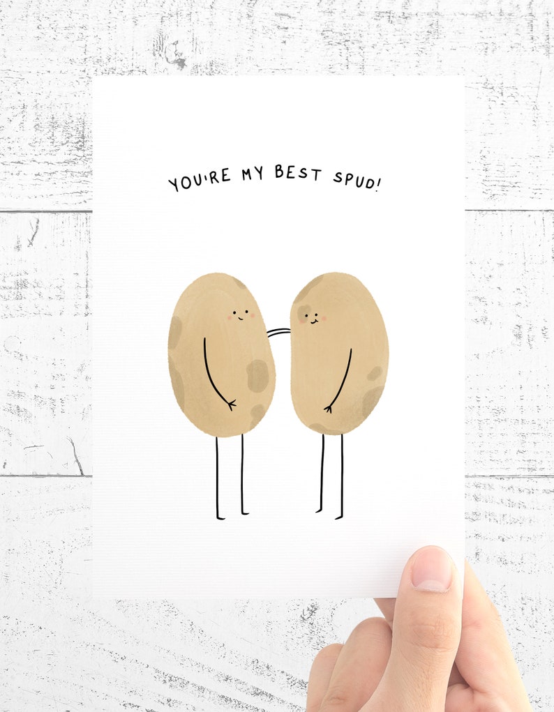 May include: A greeting card with the text "YOU'RE MY BEST SPUD!" features two cartoon potatoes with arms and legs. The potatoes are light brown with darker spots and are smiling. The card is held by a hand.