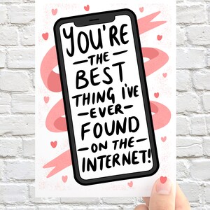 Op de afbeelding: Een wenskaart met een smartphone-illustratie en de tekst "You're the best thing I've ever found on the internet!" in het zwart. De kaart heeft een witte achtergrond met roze hartjes en linten.