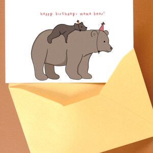 Pu&ograve; includere: Un biglietto di compleanno con un'illustrazione a cartoni animati di un grande orso marrone con un orso pi&ugrave; piccolo sulla schiena, entrambi con cappelli da festa. Il biglietto dice "happy birthday, mama bear!" ed &egrave; in una busta gialla.