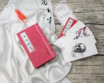 Puede incluir: Una caja roja con caracteres chinos en la parte delantera, una bufanda de seda blanca con caracteres chinos y un juego de cartas con ilustraciones de cráneos en blanco y negro. Las cartas tienen caracteres chinos y el texto "Oriental Skull".