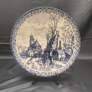 Op de afbeelding: Een ronde keramische bord met een blauw-witte transferprint van een winters dorpsgezicht met een kerk, kale bomen en figuren. De rand is versierd met een bloemmotief en de tekst "De Winter" staat onderaan in het midden afgedrukt.