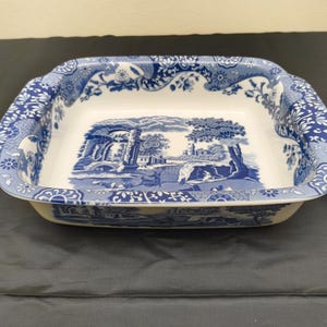 Puede incluir: Una fuente de horno cuadrada de cerámica con base blanca y un patrón azul de estilo transferware. El interior y el borde muestran ilustraciones detalladas de ruinas clásicas, árboles y paisajes, complementadas con intrincados bordes florales.