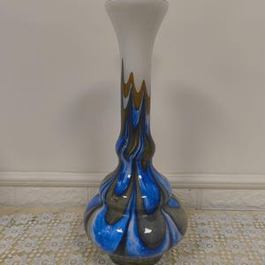 Könnte beinhalten: Eine hohe, dekorative Glasvase mit ausgestelltem Hals und bauchigem Boden. Die Vase hat einen weißen oberen Teil, der in ein Design aus blauen, grauen und goldenen Wirbeln übergeht. Die Vase ist etwa 36 cm hoch.