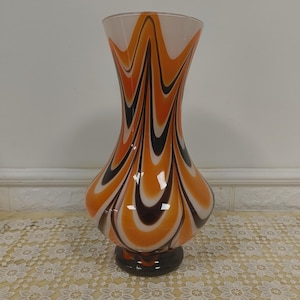 Könnte beinhalten: Eine Vintage-Glasvase mit ausgestelltem Oberteil und bauchigem Boden. Die Vase hat ein auffälliges Design aus orangefarbenen, schwarzen und weißen Wirbelmustern. Der Sockel ist dunkelbraun und setzt einen Kontrast. Ein dekoratives Stück.