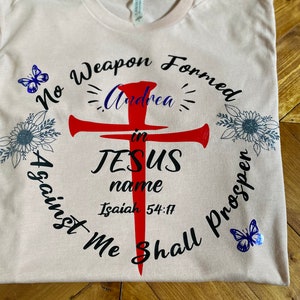 May include: A pink t-shirt with a black and red cross design. The text "No Weapon Formed Against Me Shall Prosper" is written around the cross. The name "Andrea" is written inside the cross. The text "in JESUS name" is written below the cross. The text "Isaiah 54:17" is written below the name. There are two blue butterflies on the shirt.