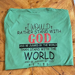 Peut inclure: T-shirt vert avec un imprimé graphique noir et rouge qui dit "Je préférerais rester avec Dieu et être jugé par le monde plutôt que de rester avec le monde et être jugé par Dieu". Le graphique comprend également cinq croix.
