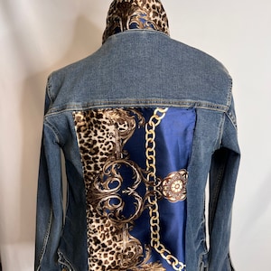 May include: A blue denim jacket with a patterned fabric lining on the back and collar. The fabric features a gold chain design, leopard print, and floral patterns.