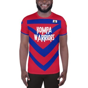Puede incluir: Camiseta roja con detalles azules y el texto blanco "KOMPA WARRIORS". La camiseta presenta un diseño geométrico con rayas de chevron azules en el pecho y los hombros. El diseño incluye la bandera haitiana.