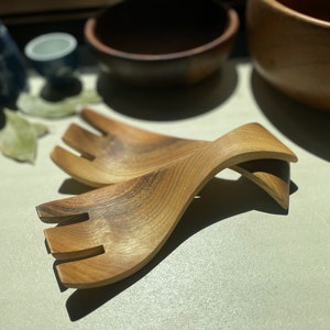 May include: A pair of wooden salad servers with a unique curved design. The servers are made of light brown wood and have a natural finish.