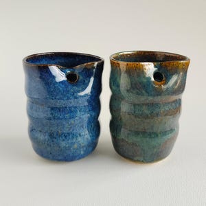 May include: Two cylindrical ceramic cups with a rippled texture and a small circular hole near the rim. One cup features a vibrant blue glaze with darker speckles, while the other displays a blend of deep teal, green, and brown tones.