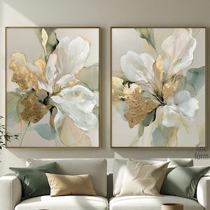 May include: Two square wall art canvases featuring abstract floral compositions in white, soft green, and metallic gold leaf accents. The paintings are displayed side-by-side above a white sofa, with the text line form visible in the lower right corner.
