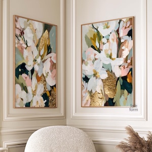 May include: Two framed abstract floral paintings with gold leaf accents. The artwork features white, pink, and green brushstrokes on a dark background. The paintings are displayed in a corner of a room with a white, rounded armchair.