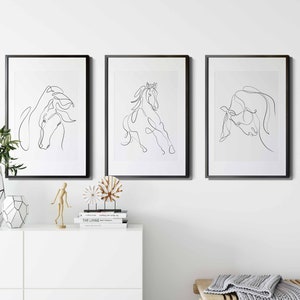 May include: Three black framed prints of line drawings of horses. The prints are hanging on a white wall above a white dresser with a wooden decorative object and a wooden mannequin. A woven basket is on the floor below the dresser. A wooden bench with a grey knitted throw is in the foreground.