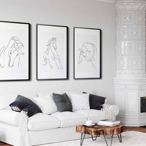 May include: Three black framed prints of line drawings of horses hanging on a white wall above a white sofa with black throw cushions. The sofa is in a living room with a white tiled fireplace and a wooden coffee table.