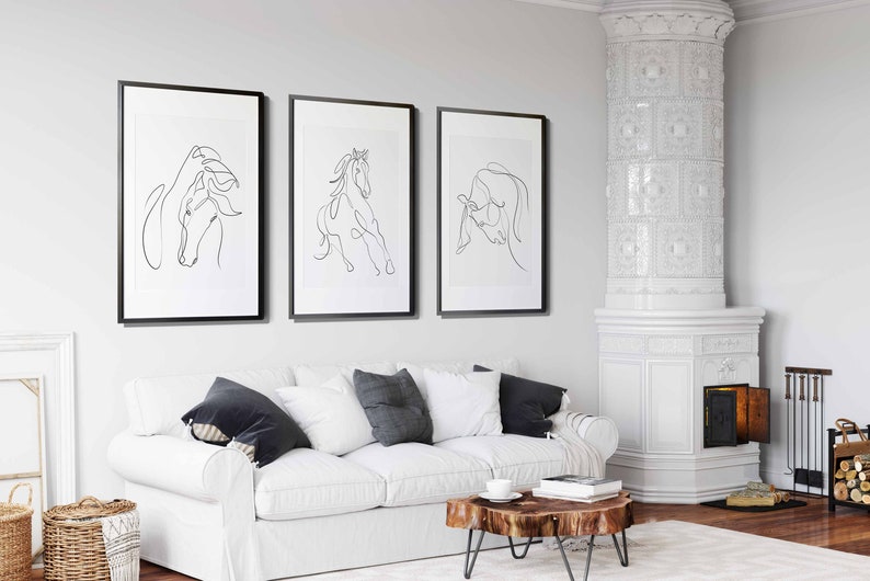 May include: Three black framed prints of line drawings of horses hanging on a white wall above a white sofa with black throw cushions. The sofa is in a living room with a white tiled fireplace and a wooden coffee table.