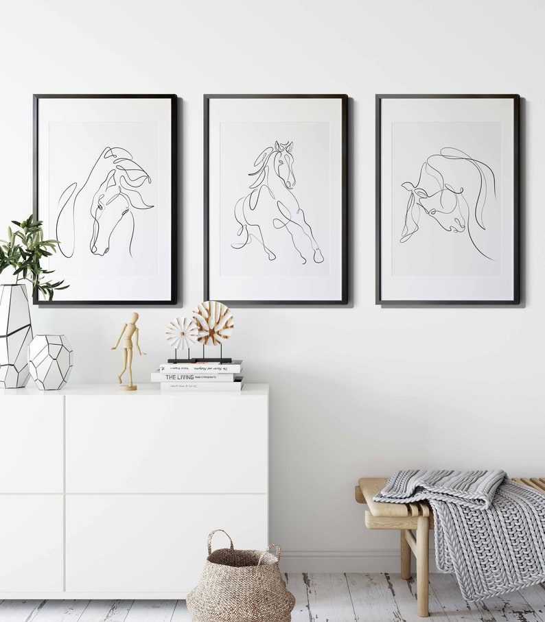 May include: Three black framed prints of line drawings of horses. The prints are hanging on a white wall above a white dresser with a wooden decorative object and a wooden mannequin. A woven basket is on the floor below the dresser. A wooden bench with a grey knitted throw is in the foreground.