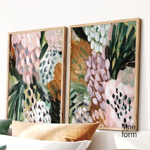 May include: Two framed abstract art prints. The artwork features a floral design with pink, green, and brown brushstrokes. The frames are made of light-colored wood. The prints are displayed on a white wall.