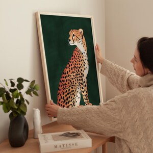 May include: A framed print of a cheetah sitting on a dark green background. The cheetah is facing to the left and has a spotted coat with orange and black markings.