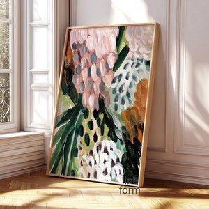 May include: Abstract floral art print in a wooden frame. The artwork features a mix of pink, green, orange, and white brushstrokes. The painting is leaning against a white wall and a window with natural light.