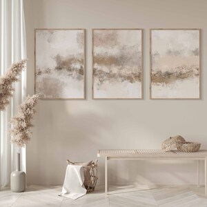 May include: Three abstract paintings in beige, brown, and white tones are hung on a light beige wall. The paintings are framed in natural wood. Two tall vases with dried pampas grass are on the left side of the image. A wooden bench with a woven basket on top is in the foreground.