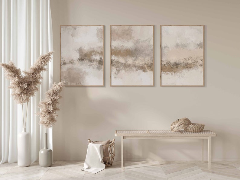 May include: Three abstract paintings in beige, brown, and white tones are hung on a light beige wall. The paintings are framed in natural wood. Two tall vases with dried pampas grass are on the left side of the image. A wooden bench with a woven basket on top is in the foreground.