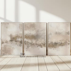 May include: Three abstract art prints in white frames with a beige and brown watercolor-like design. The prints are hung on a white wall with a wood floor.