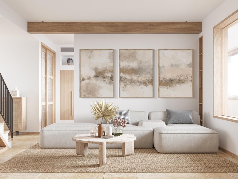 May include: A white sectional sofa with three cushions, a coffee table, and three abstract paintings on the wall. The sofa is in a living room with a wooden beam ceiling and a large window.