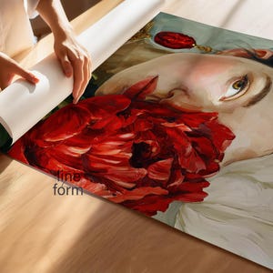 Puede incluir: Una persona desenrolla una impresi&oacute;n art&iacute;stica que presenta un retrato de una persona con una gran flor roja. La obra de arte tiene un estilo pict&oacute;rico, con un enfoque en el rostro y el rojo vibrante de la flor. La impresi&oacute;n est&aacute; sobre una superficie de madera.