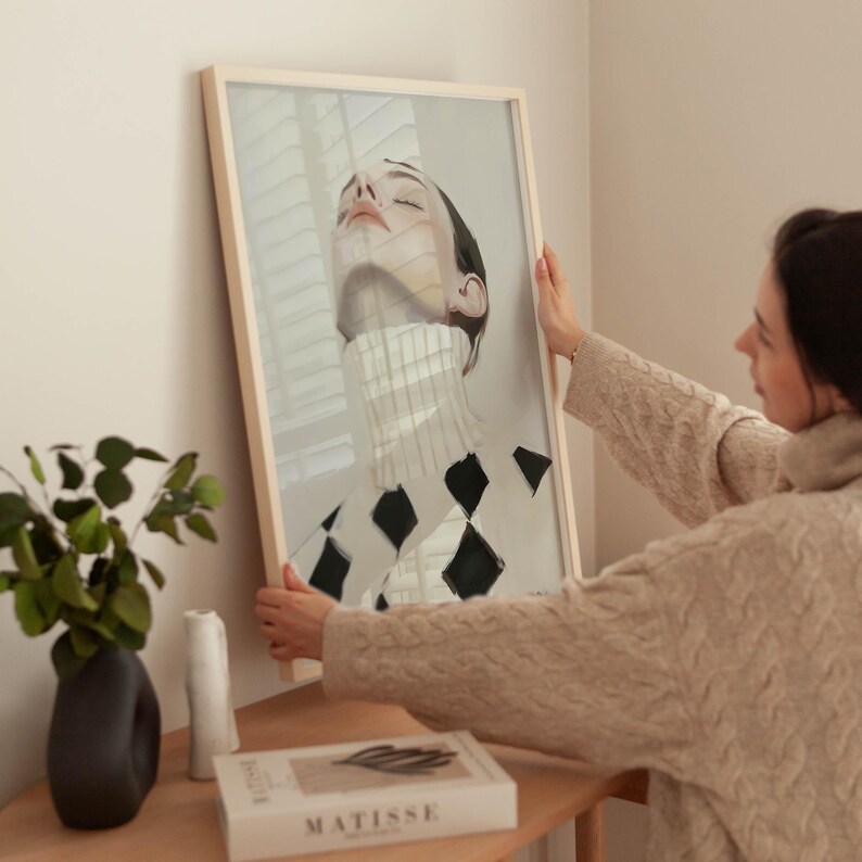 Puede incluir: Una impresi&oacute;n art&iacute;stica enmarcada que presenta a una mujer con los ojos cerrados, con un cuello alto blanco y un patr&oacute;n de diamantes en blanco y negro. La obra de arte es sostenida por una persona con un su&eacute;ter beige. La impresi&oacute;n est&aacute; en un marco de madera clara.