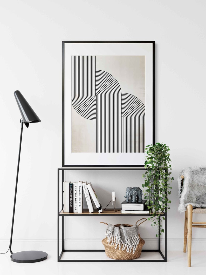 May include: A black framed print with a geometric design of overlapping black lines on a beige background. The print is displayed on a black metal shelf with books and a small elephant figurine.