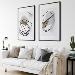 May include: Two black framed abstract art prints with white backgrounds and black lines. The prints are hanging on a white wall above a white couch with pillows and a throw blanket.