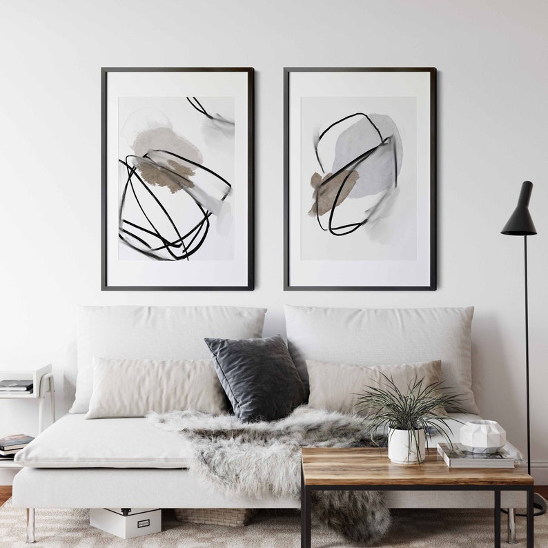 May include: Two framed abstract art prints with black lines and watercolor washes in shades of gray and brown. The prints are hanging on a white wall above a white couch with gray and beige pillows.