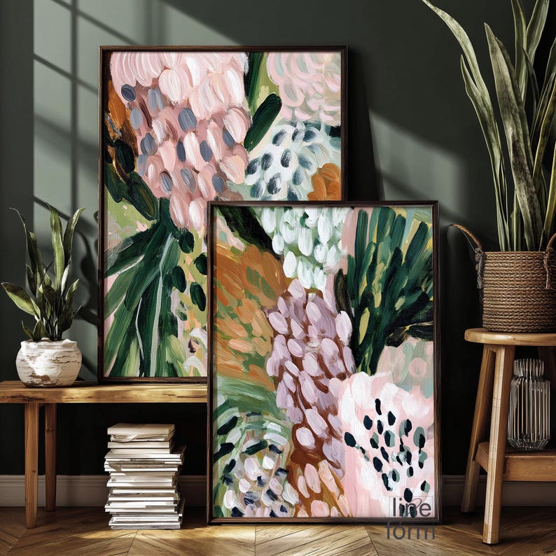 May include: Two framed abstract floral paintings. The artwork features a mix of pink, green, white, and brown brushstrokes. The paintings are displayed on a wooden surface, with a stack of books and potted plants nearby.