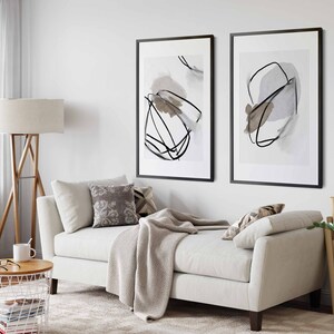 May include: A white couch with a throw blanket and pillows in a living room with two black framed abstract art prints on the wall, a wooden floor, a floor lamp, a side table, a potted plant, and a window with white curtains.