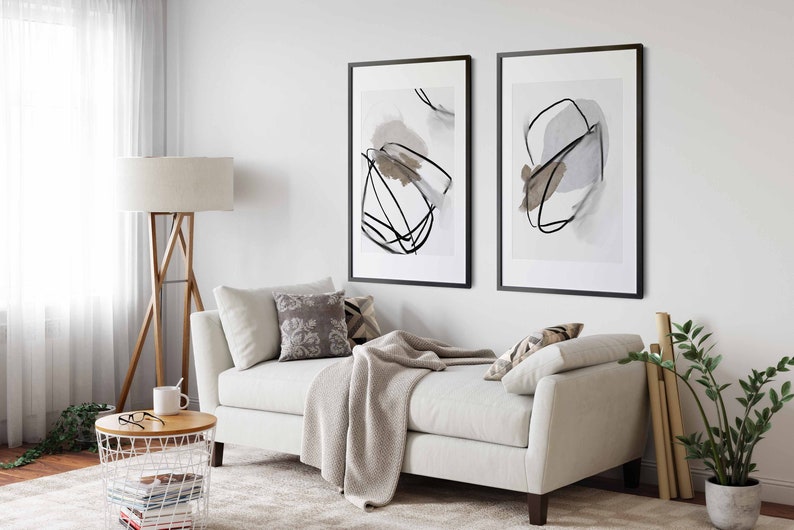 May include: A white couch with a throw blanket and pillows in a living room with two black framed abstract art prints on the wall, a wooden floor, a floor lamp, a side table, a potted plant, and a window with white curtains.