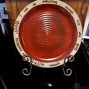 Könnte beinhalten: Ein runder Keramikteller mit einer tiefroten, spiralförmigen Mitte und einem cremefarbenen Rand. Der Rand ist mit dem schwarzen Text "I can do everything through Him who gives me strength" und "Philippians 4:13" sowie kleinen blattartigen Verzierungen versehen.