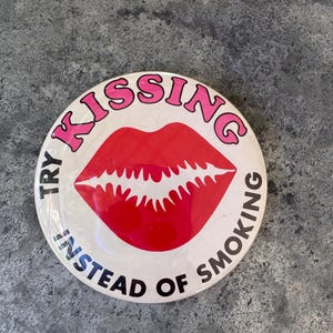 Może przedstawiać: Okrągły biały przypinany znaczek z centralną grafiką jaskrawoczerwonych ust. Napis "TRY KISSING INSTEAD OF SMOKING" jest nadrukowany w okrągły wzór wokół ust, przy czym "KISSING" jest różowe, a "TRY" oraz "INSTEAD OF SMOKING" są czarne.