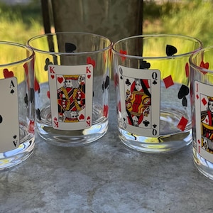 May include: Four clear drinking glasses with playing card designs. Each glass features a different card: Ace of Spades, King of Hearts, Queen of Hearts, and Jack of Diamonds. Red and black suit symbols decorate the glass.