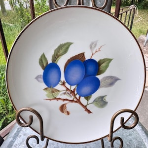 May include: A white ceramic plate with a brown rim and a decorative plum design. The design features several blue plums with green leaves and brown branches. The plate is displayed on a decorative metal stand.