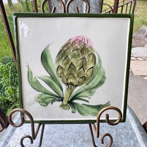 May include: Square ceramic tile with a hand-painted artichoke design. The artichoke is green and pink, with green leaves, set against a white background. The tile has a green border and is displayed on a decorative metal stand.