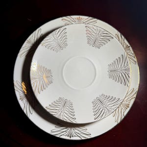 Puede incluir: Juego de vajilla blanco y dorado de dos piezas. El juego incluye un platillo y un plato más pequeño, ambos con un diseño de hojas doradas. Los bordes de los platos también están adornados con dorado. Los platos están apilados.