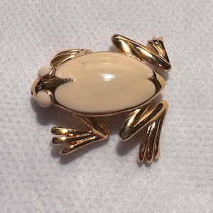 May include: A gold-toned frog brooch with a cream-colored body and white accents. The frog's legs and details are intricately designed, creating a charming and detailed accessory. This vintage-style brooch is a unique piece of jewelry.