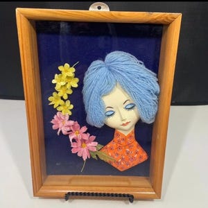 May include: Framed artwork featuring a ceramic face with blue yarn hair and an orange patterned top. Yellow and pink flowers are arranged on the left side of the face, set against a dark blue background. The artwork is encased in a wooden frame.